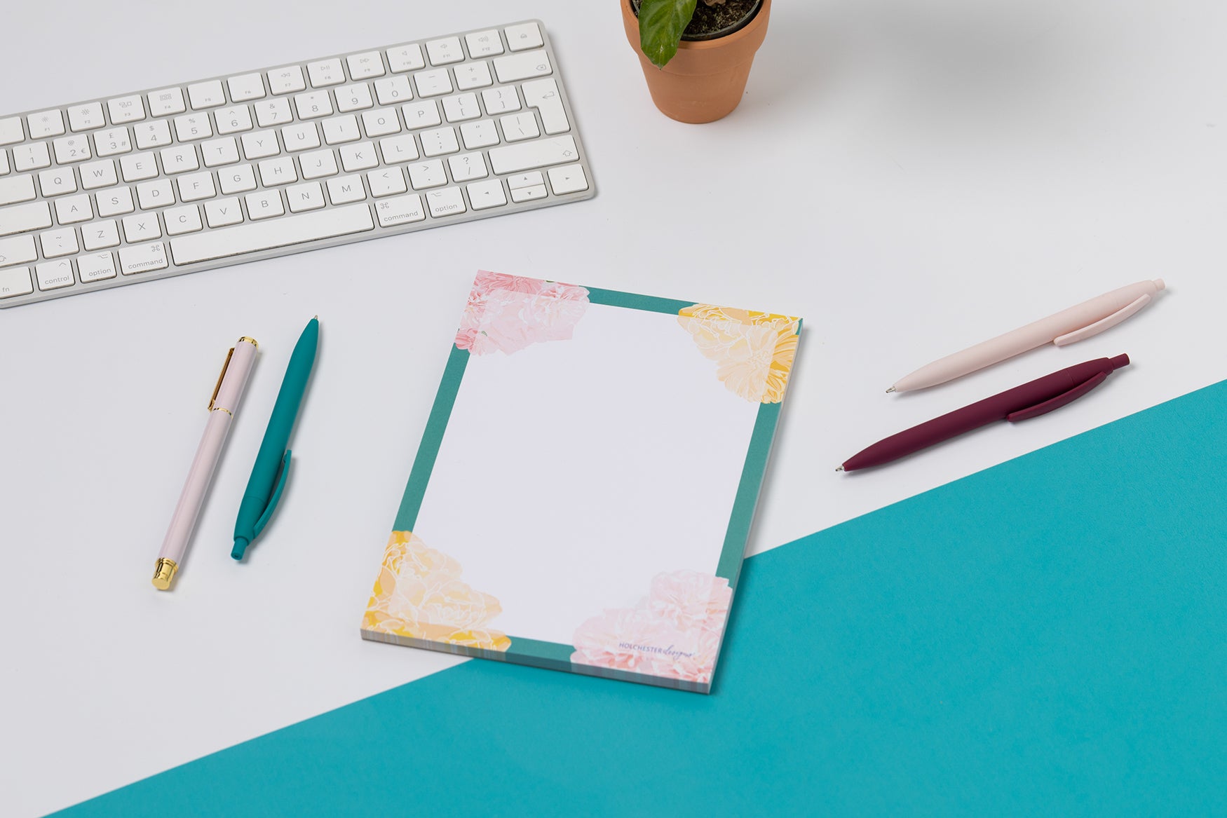Spring Forward A5 Notepad is flat on a tabletop - half teal and half white cutting across the image diagonally.  There pens and white keyboard around it.