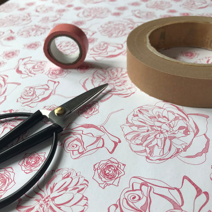 Spring Forward wrapping paper - white background with dark pink line-drawn floral motifs - is flat on a table, with a small pair of scissors, small roll of pink tape and a larger roll of brown tape lying on it.