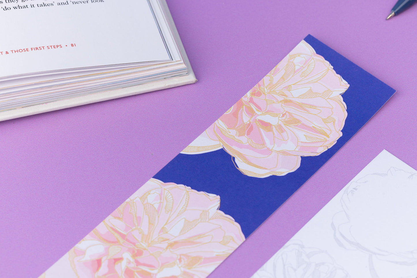 Close up of the Spring Forward Bookmark with a blue background and pink dahlia illustrations and golden yellow outlines.  Next to is is an out of focus corner of the bookmark's other side. They are on a lilac desk with an open book in the top left corner.