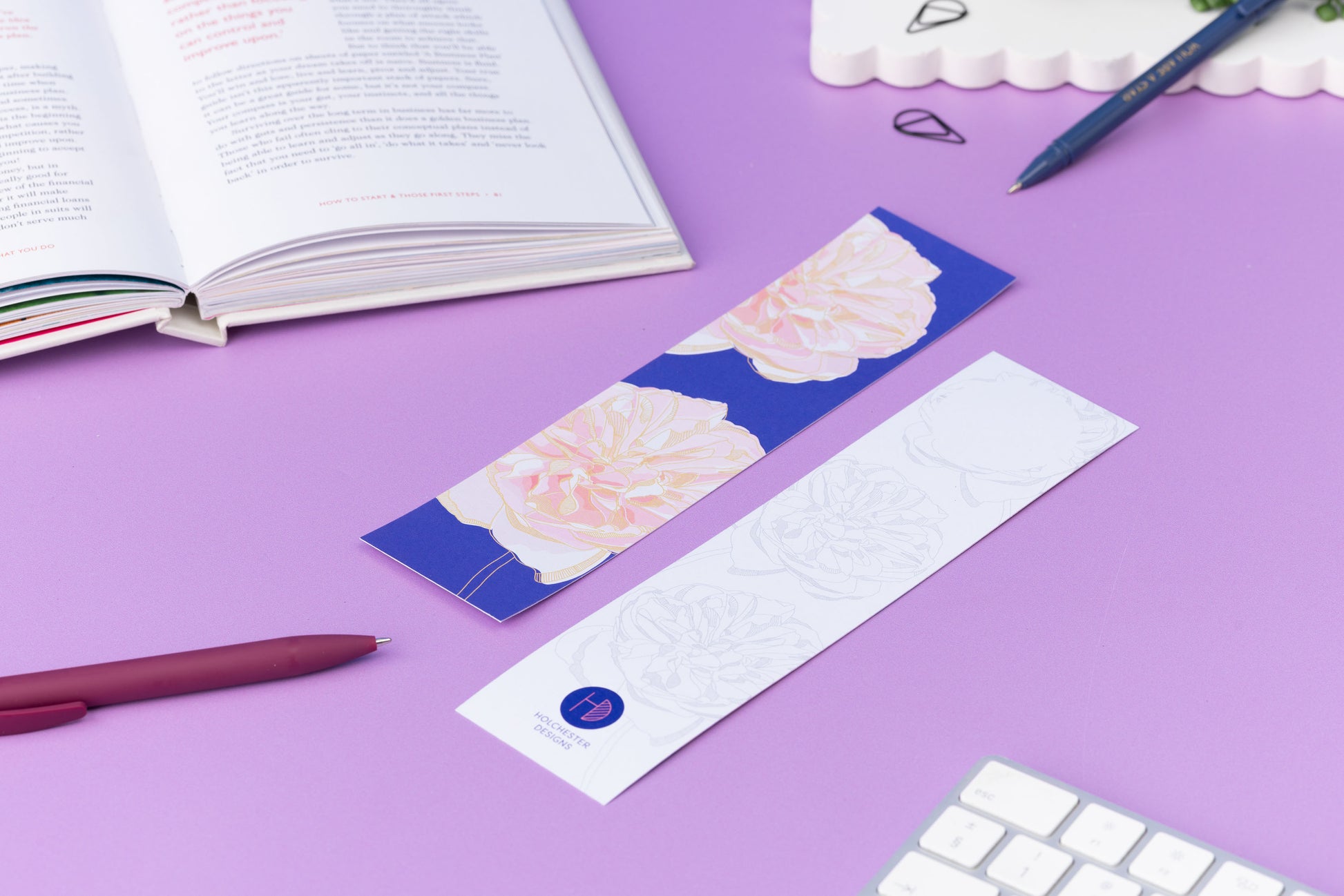 Two bookmarks, showing each side of the Spring Forward Bookmark.  One side has a blue background with pink dhalia illustrations and golden ellow outlines, the other side is white with light grey outlines of the dahlia.  They are on a lilac desk with an open book, pens and keyboard around them.