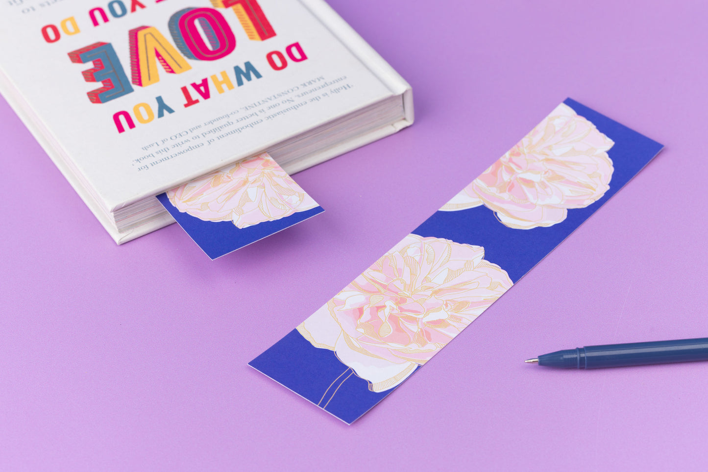 Spring Forward double-sided bookmark is lying, full colour pattern up, on a lilac desk.  To its left is a closed book with the same side of the bookmark sticking out of it.
