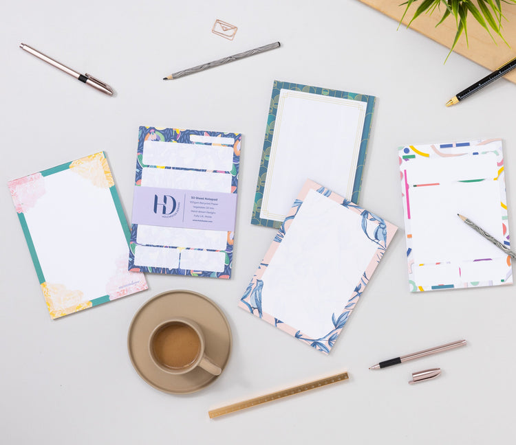 4 A5 notepads and 1 A6 Notepad from Holchester Designs are scattered on a teal desk, with pens, a small plant, a white keyboard and paperclips dotted around them.