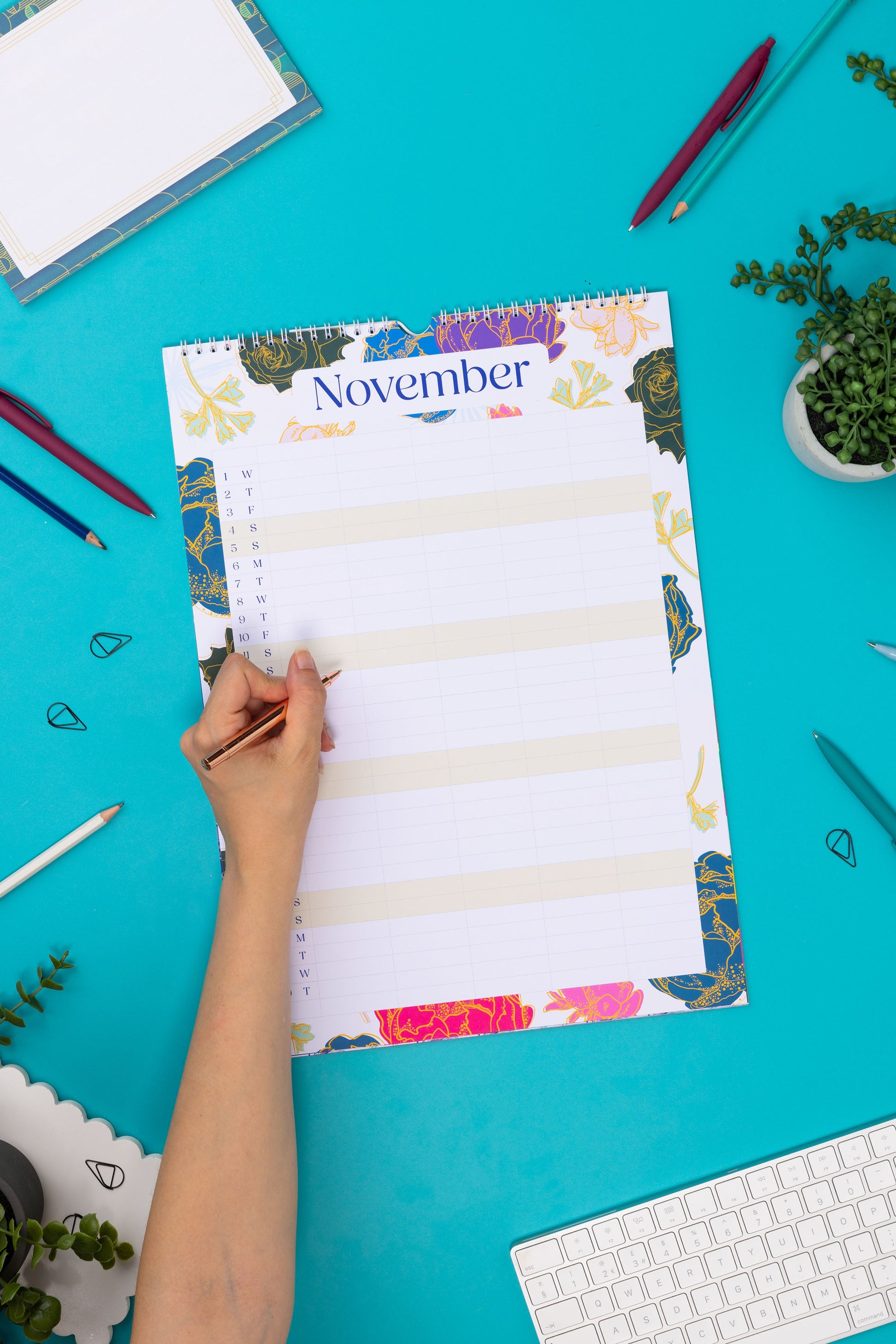 November page of the A3 sized 2024 Household Calendar.  It is lying flat on a teal desk with pens and paperclips scattered around it and a hand holding a pen to its left ready to write on it.