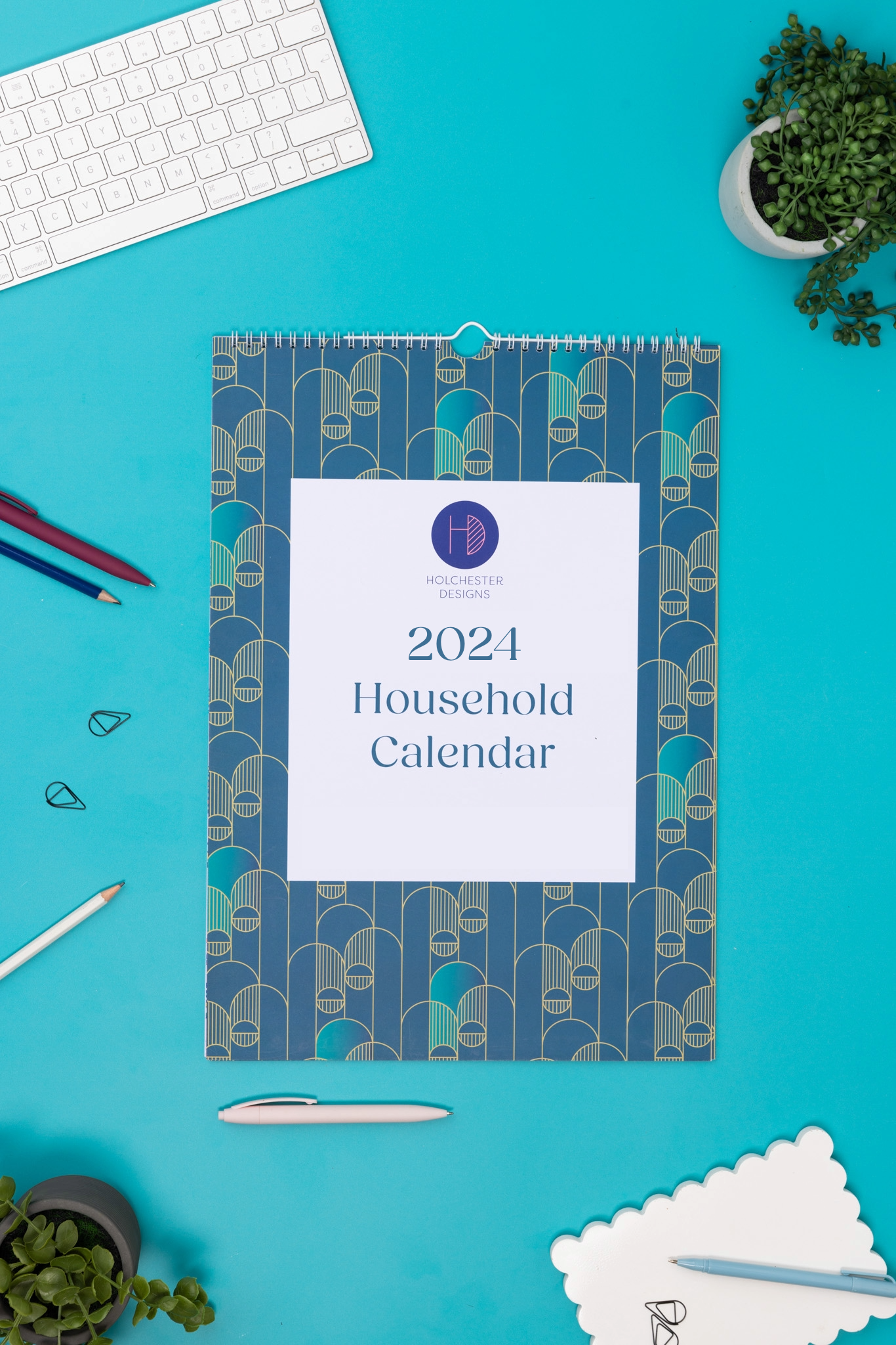 Front cover of the A3 sixed 2024 Household Calendar.  It is lying flat on a teal desk with pens and paperclips scattered around it.