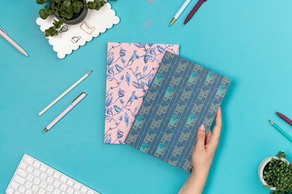 The Pair of B5 dot grid notebooks - Brighton Birds below and Deco Delights being held by a hand to its right.  They are on a teal desk with a white keyboard, two small plants and scattered pens around them.