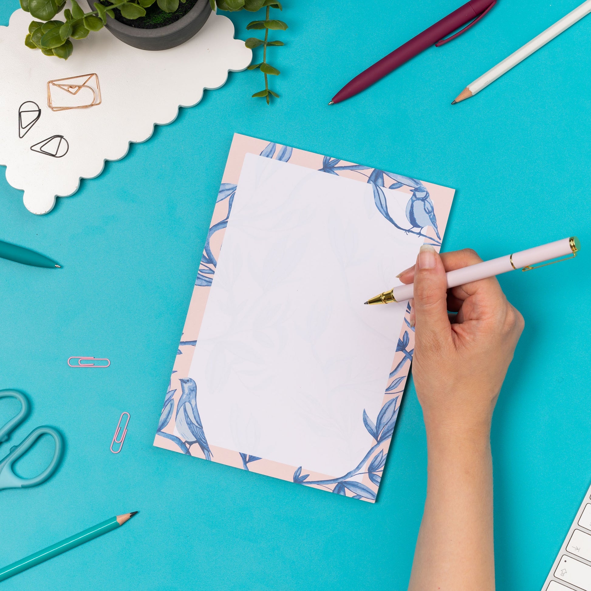 Brighton Birds A5 Notepad - Blush pink background with shades of blue bird and plant hand-painted motifs - is on a teal desk, next to a white pen, white keyboard and scattered paperclips. There is a hand to its right holding a white pen ready to write on the pad.