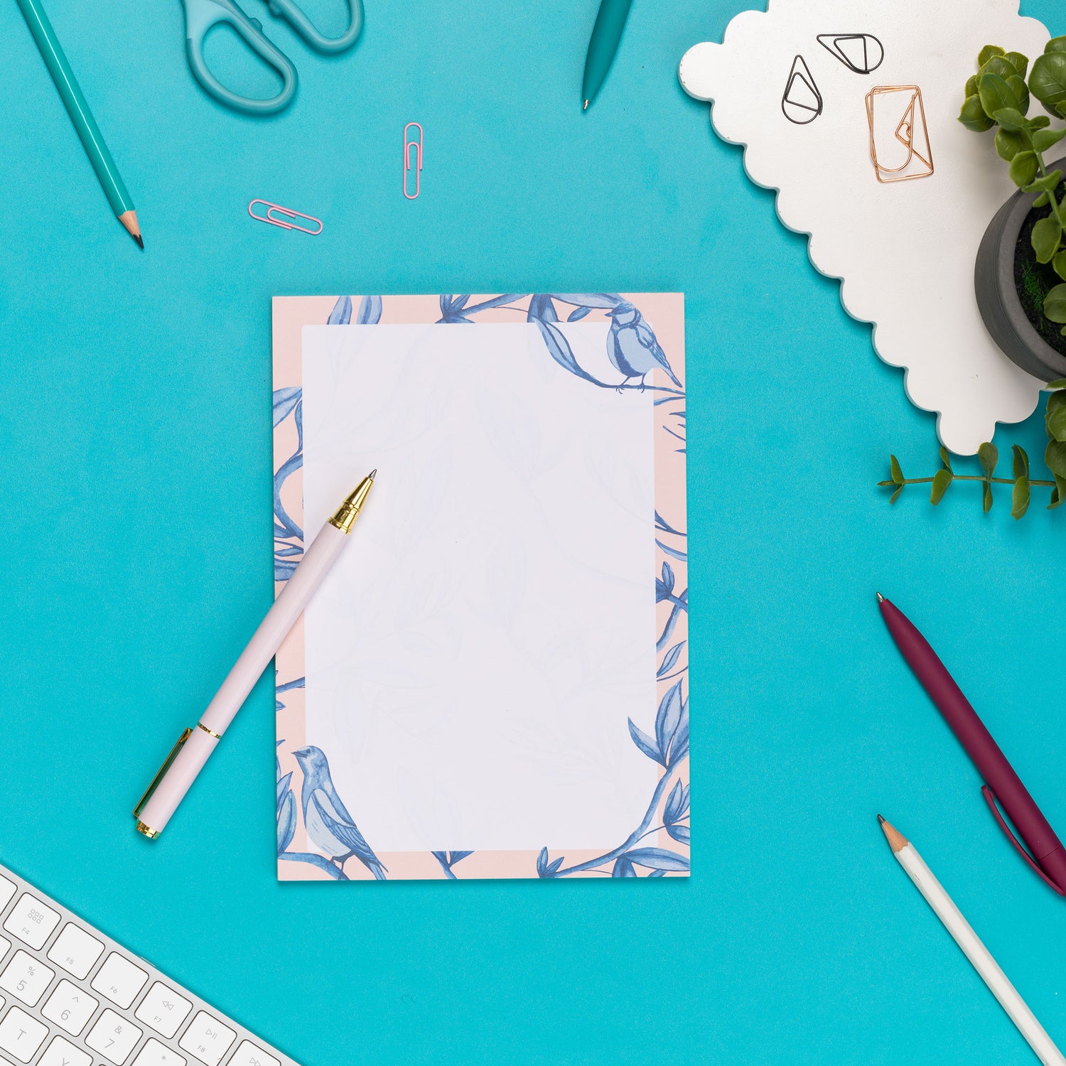 Brighton Birds A5 Notepad - Blush pink background with shades of blue bird and plant hand-painted motifs - is on a teal desk, next to a white pen, white keyboard and scattered paperclips.