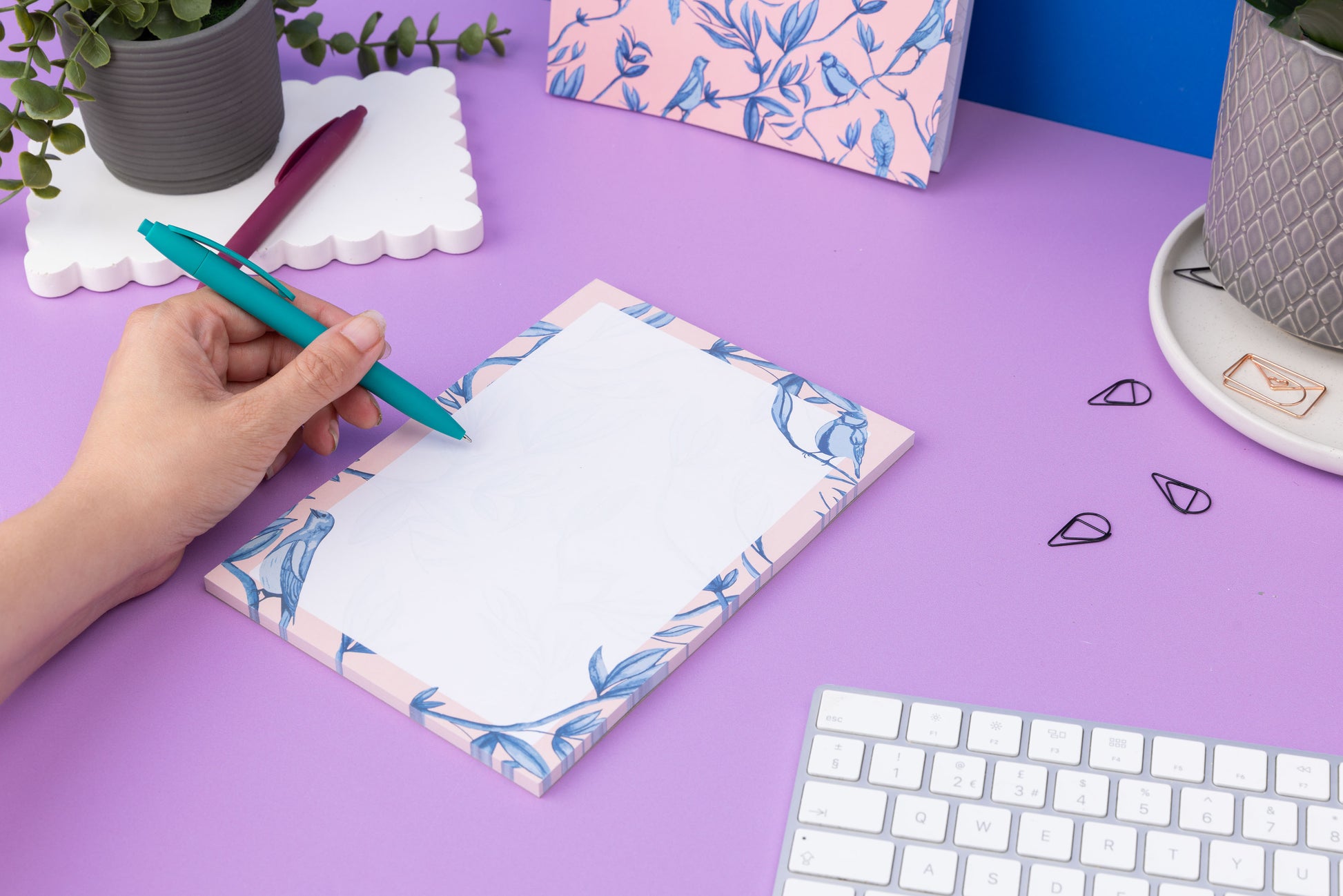 Brighton Birds A5 Notepad - Blush pink background with shades of blue bird and plant hand-painted motifs - is on a lilac desk, next to a white keyboard and scattered paperclips. There is a hand to its left holding a teal pen ready to write on the pad.