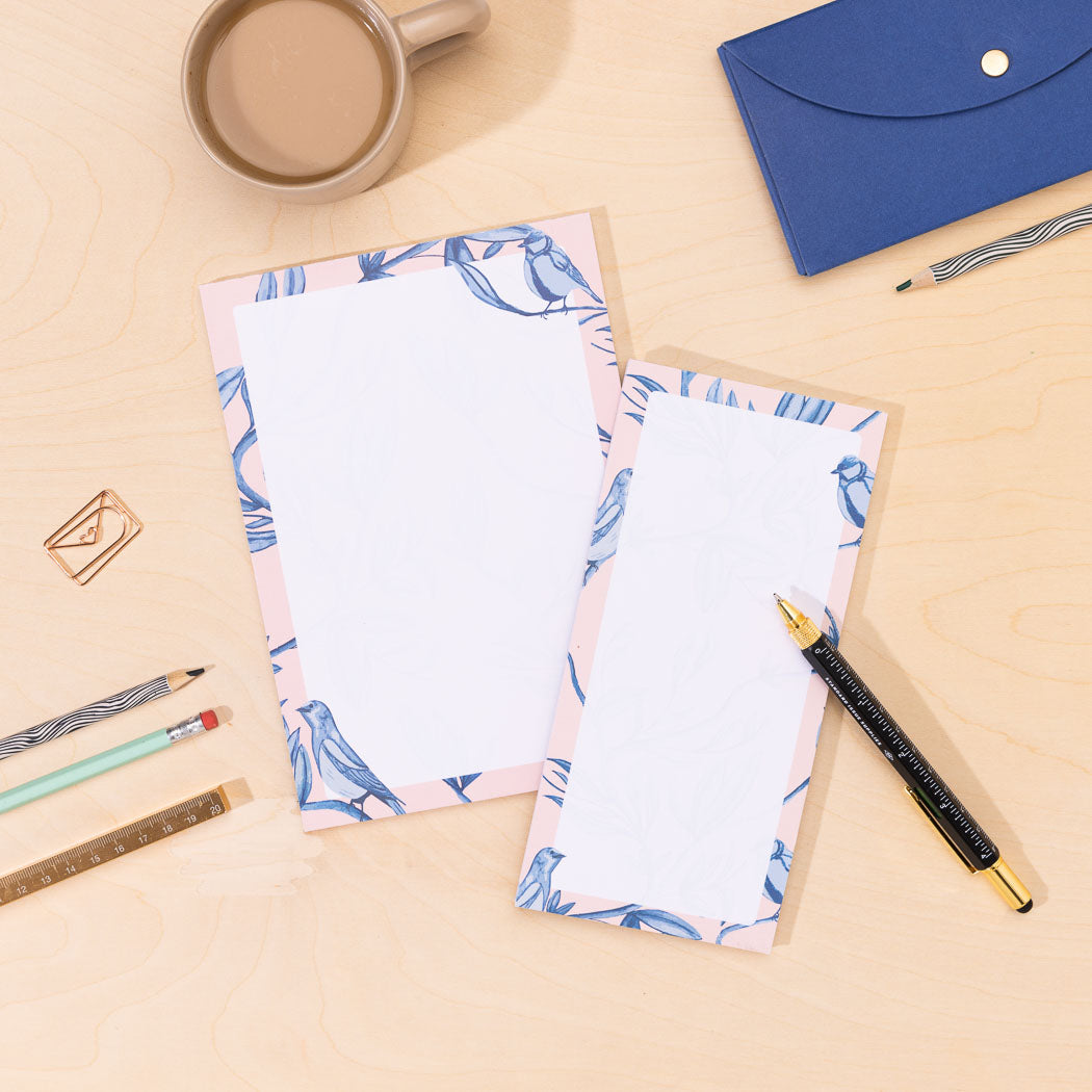 Two notepads of paper, one A5 and one DL List pad, with blue and pink border of birds and leaves on a desk with stationery items.