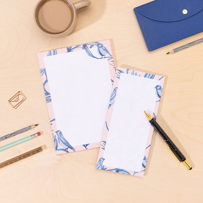 Two notepads of paper, one A5 and one DL List pad, with blue and pink border of birds and leaves on a desk with stationery items.