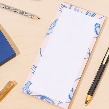 A DL list pad of paper with a blue and pink border of birds and leaves on a desk with stationery items.