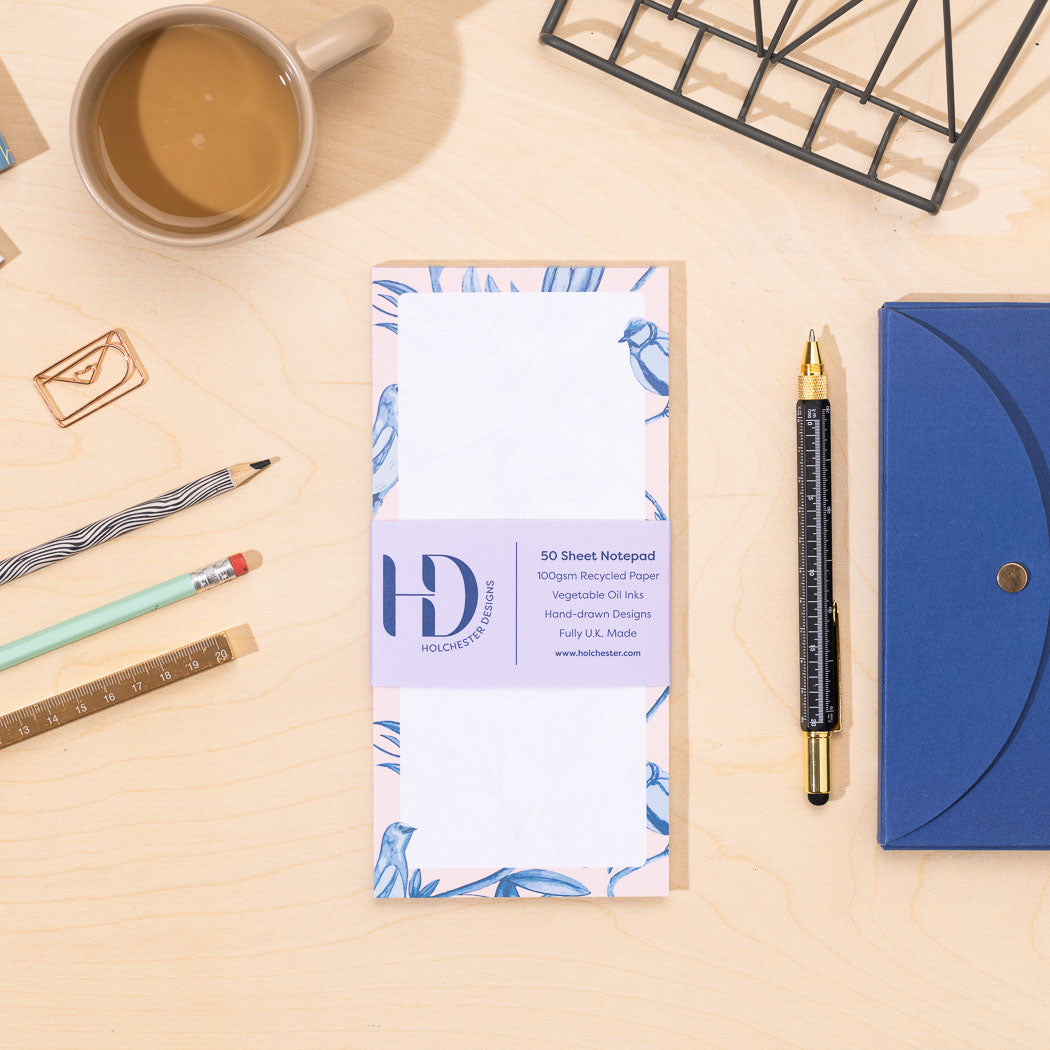A DL list pad of paper with a blue and pink border of birds and leaves on a desk with stationery items.