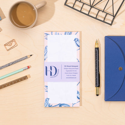 A DL list pad of paper with a blue and pink border of birds and leaves on a desk with stationery items.