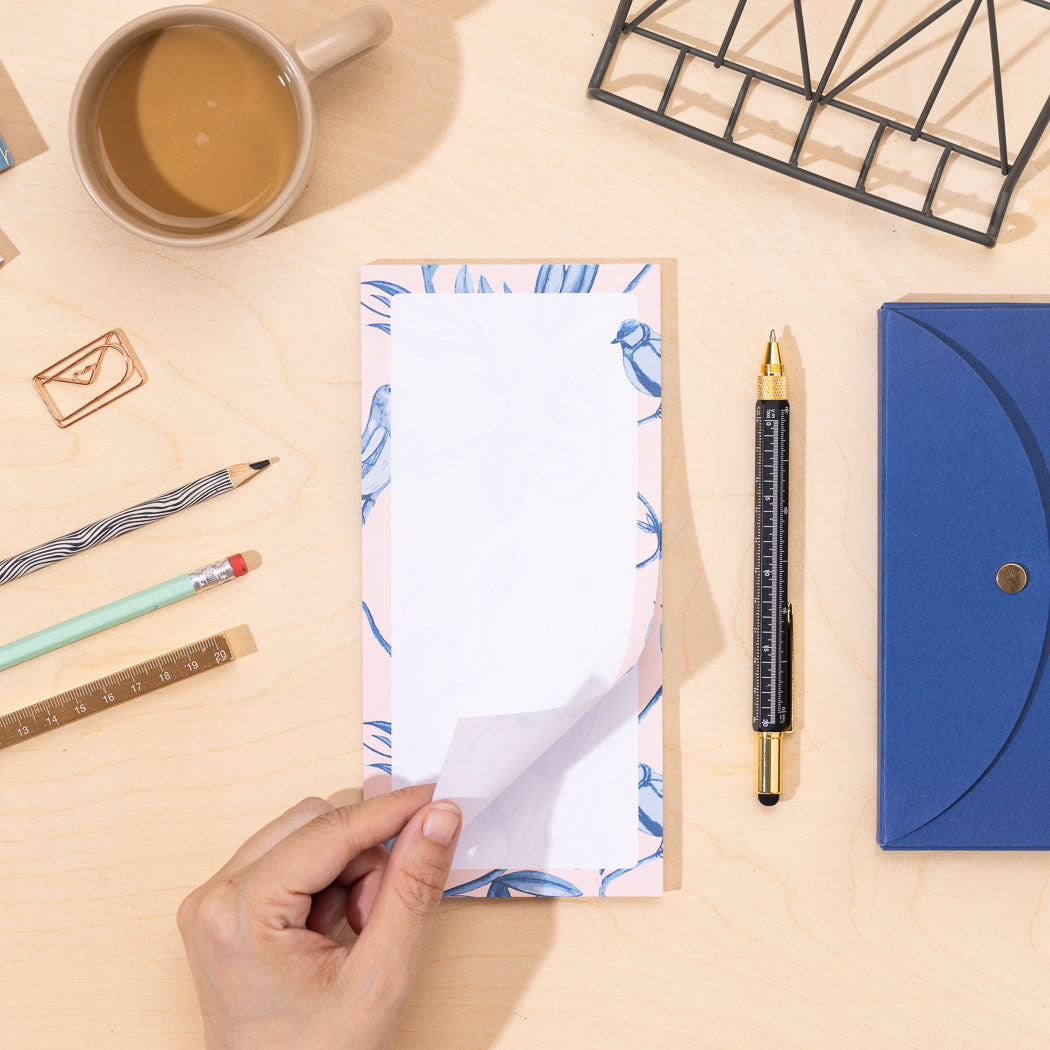 A DL list pad of paper with a blue and pink border of birds and leaves on a desk with stationery items.  A hand is turning the top page from the bottom right corner.