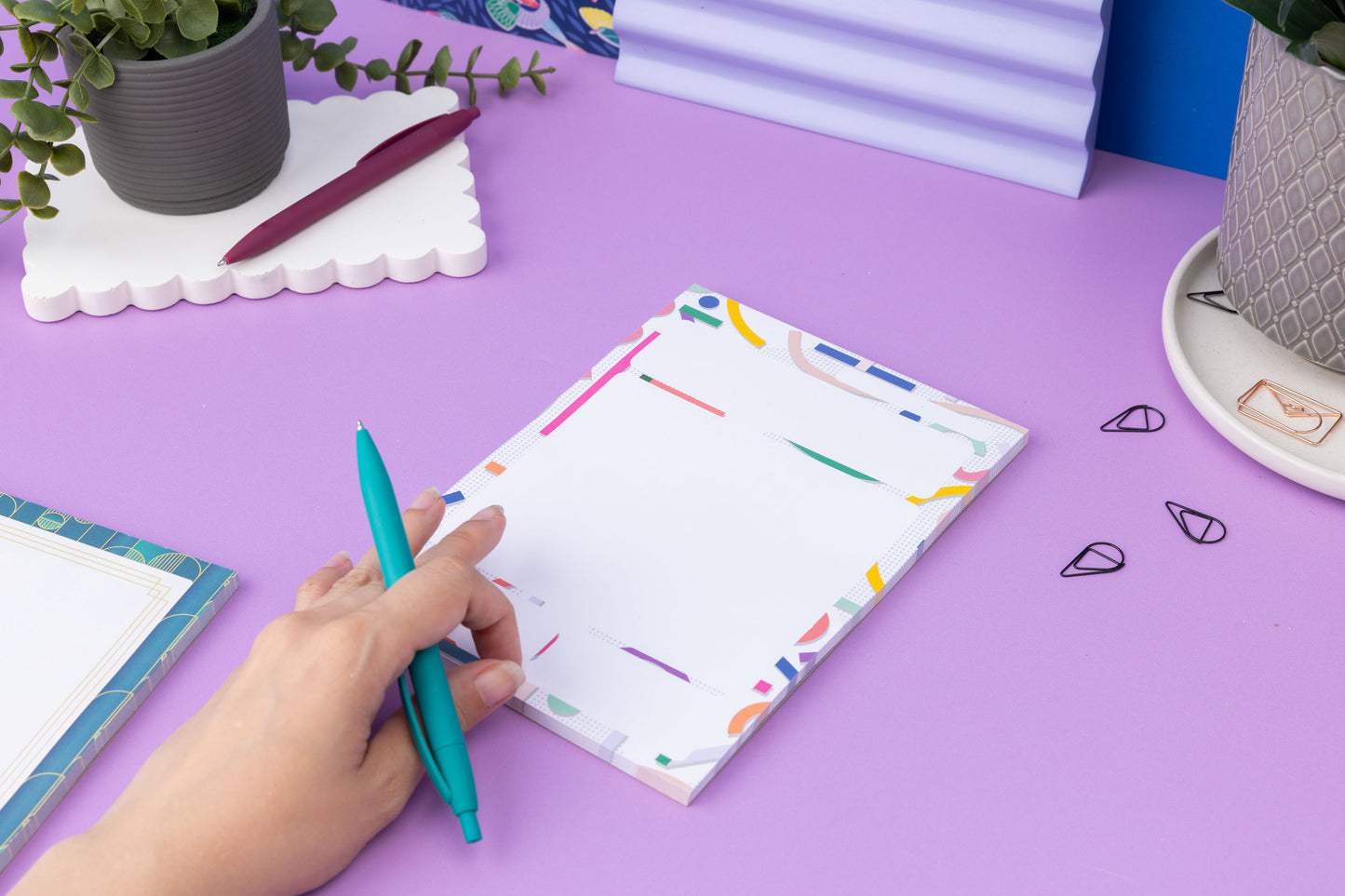 Cutouts A5 Daily Planner Pad on a lilac desk, with a hand to the bottom left holding a teal pen ready to write on it. 