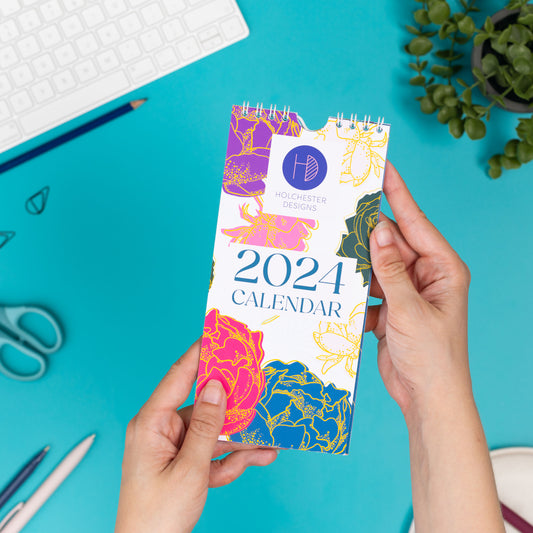 Front cover of the smaller DL 2024 calendar is being held up by two hands on its top right and bottom left.  Below it you can see, out of focus, a teal desk.