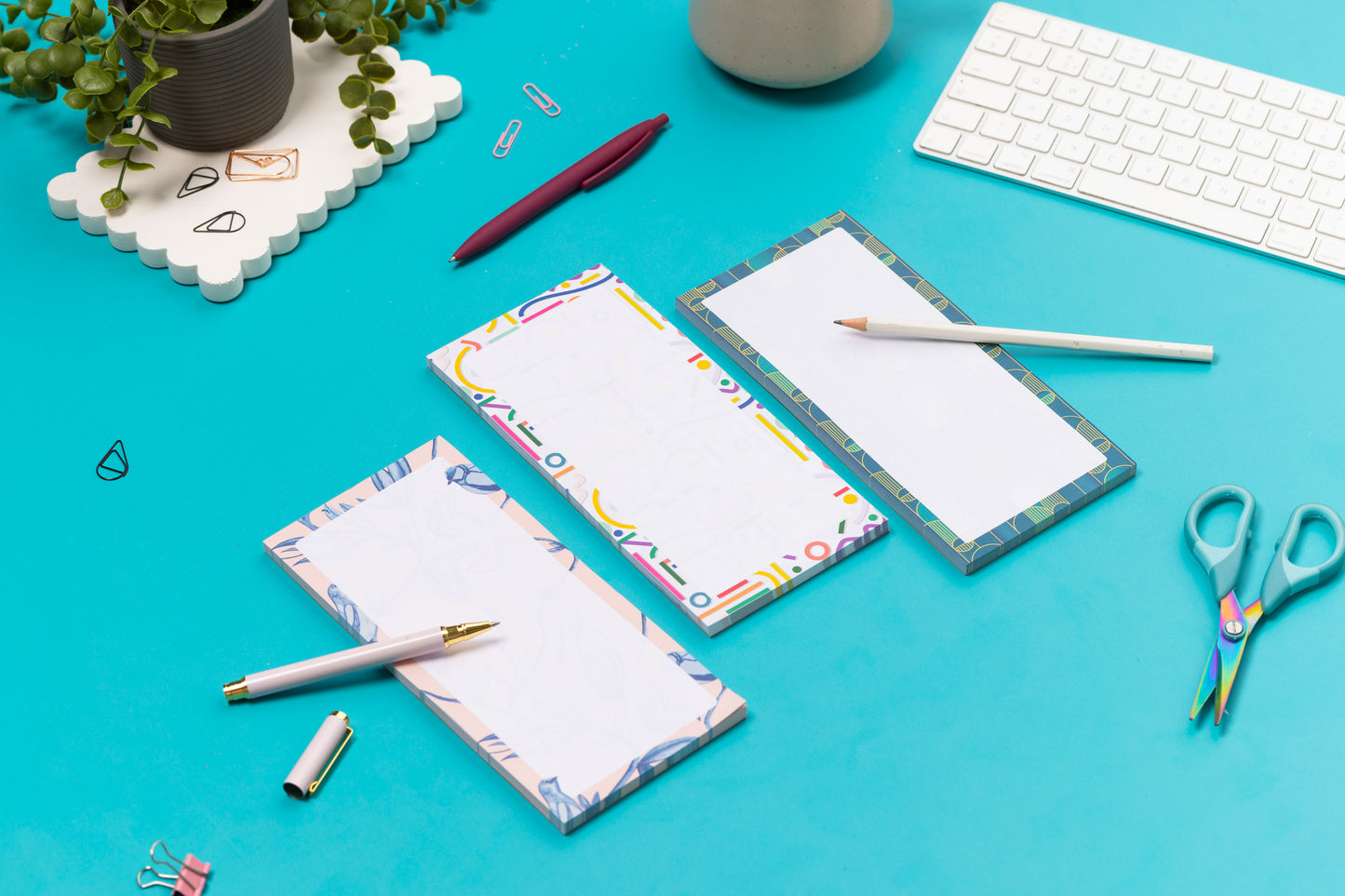 Three list pads - Brighton Birds, Cutouts and Deco Delights - are in a row on a teal desk, with a white pen and pencil leaning on them, with a white keyboard and paperclips around them.