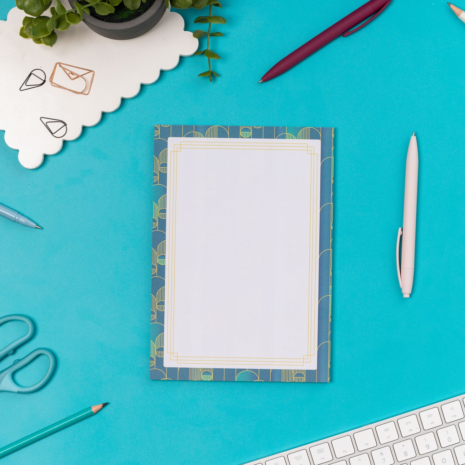 Deco Delights A5 Notepad is on a teal desk with pens and paperclips scattered around it.  There is also a white keyboard to the bottom right and small plant to the top left of the image.