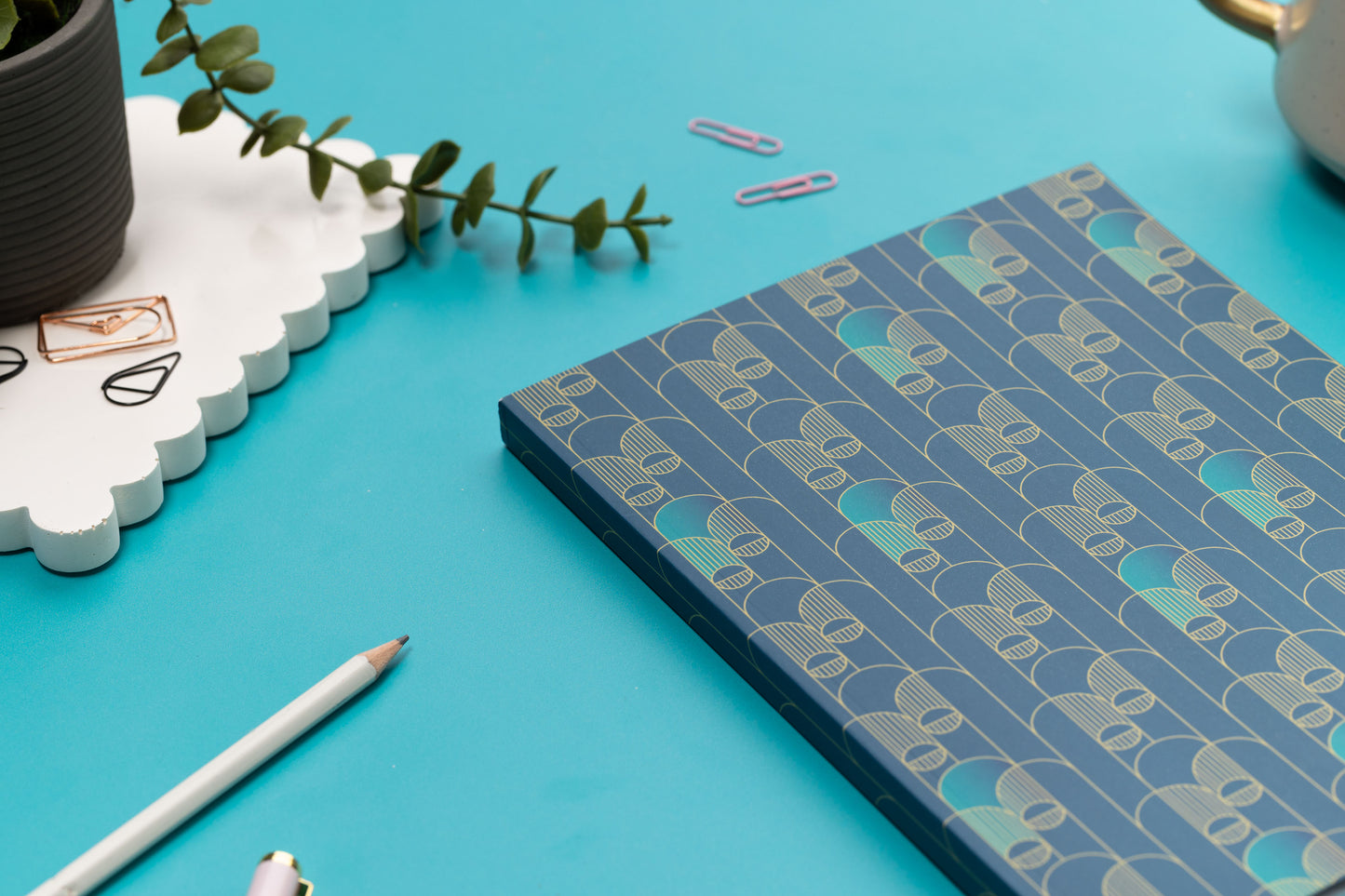 The Deco Delights B5 Notebook is facing up on a teal desk with its spine facing you to see its depth. 