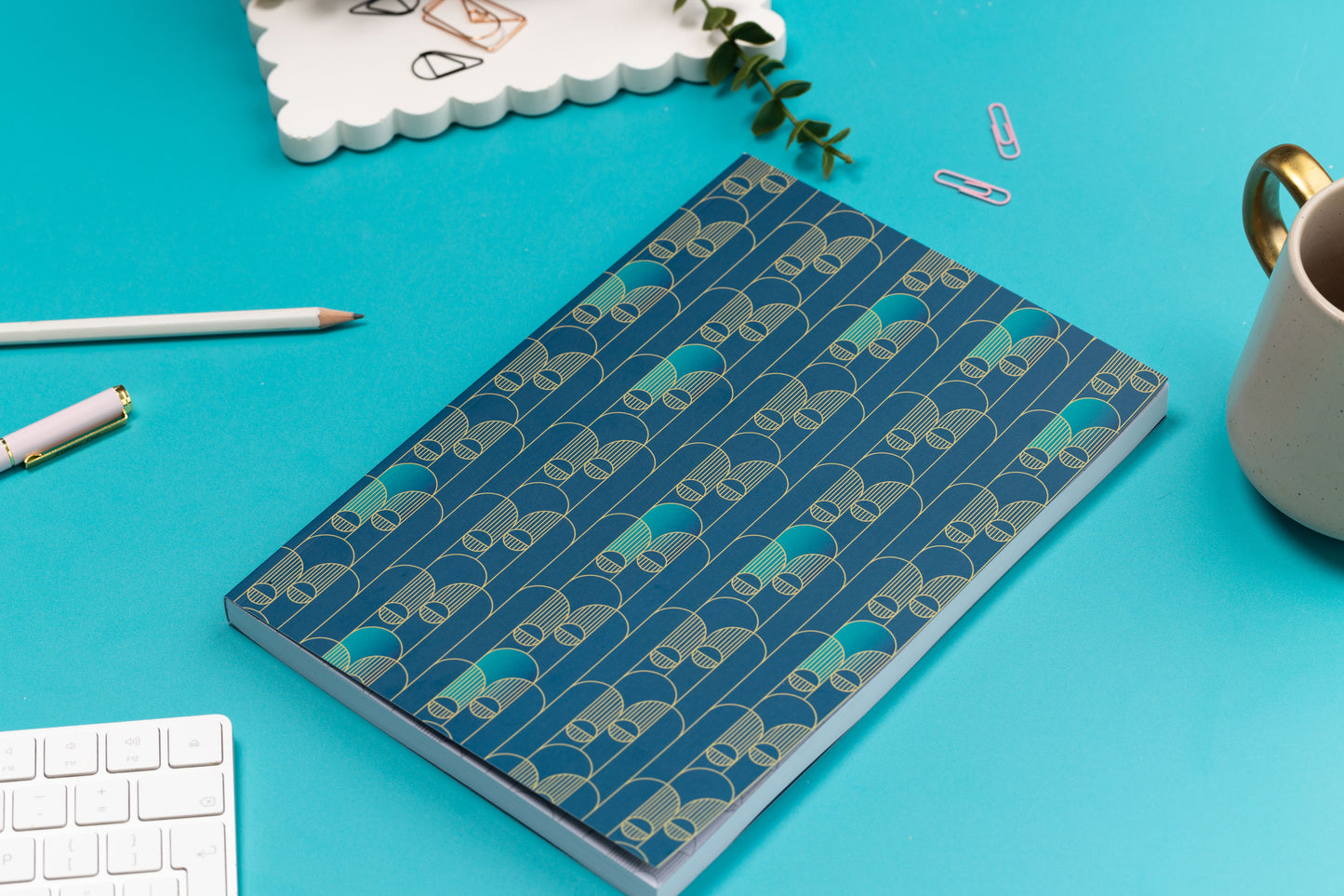 The Deco Delights B5 Notebook is facing up on a teal desk with pens and paperclips scattered around it, a white keyboard and a mug. 