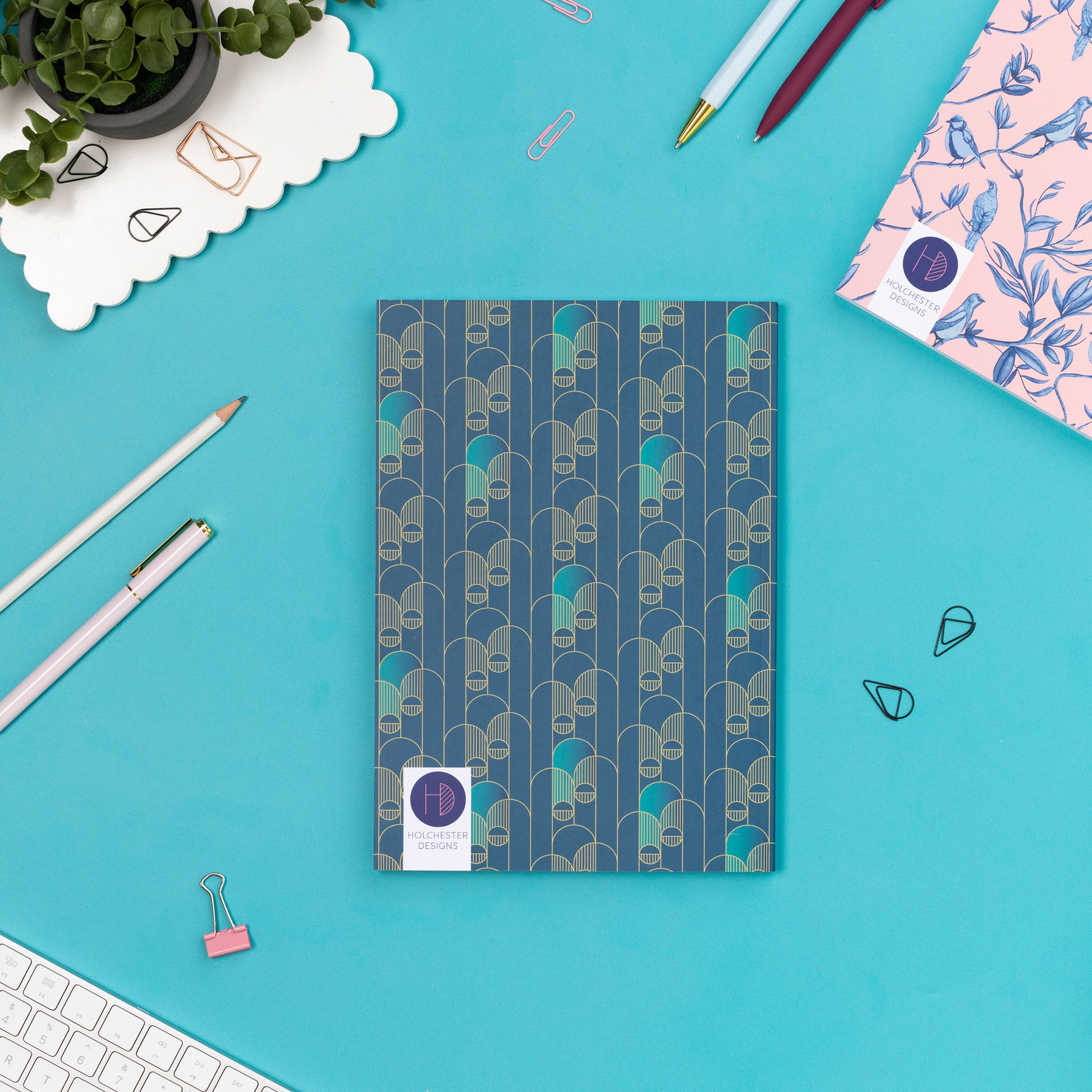 The back of the Deco Delights B5 Notebook is facing up on a teal desk with pens and paperclips scattered around it.  There is also a white keyboard to the bottom left, a small plant to the top left of the image and the bottom corner of a Brighton Birds dot grid notebook appears in the top right corner.