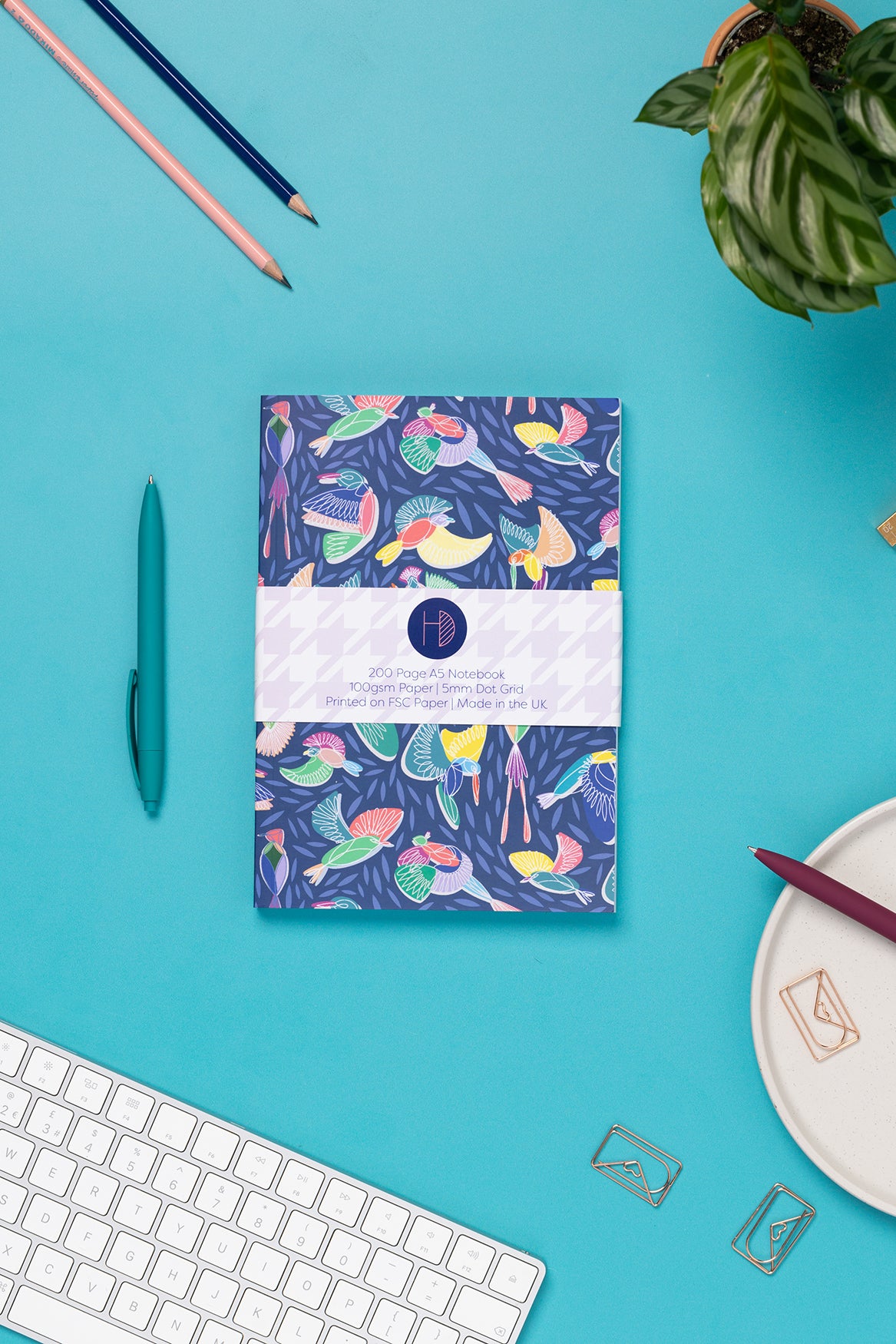 Paradise A5 Dot Grid Notebook, with its belly-band packaging around it, is on a teal desk with some pens, and a keyboard to the bottom left corner.  The book's cover features a navy background with line-drawn bird motifs and offset coloured blocks behind them.