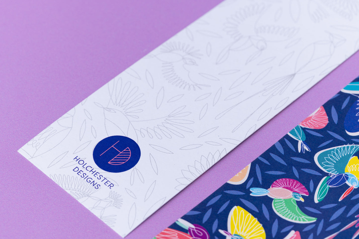 Two of the paradise double-sided bookmarks, on a lilac desk. The main focus is on the bottom of the monochrome side of the bookmark, with the Holchester Designs Logo.  The full-pattern side of the bookmark is in the bottom right corner slightly out of focus.