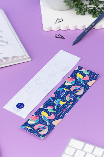 Two of the paradise double-sided bookmarks, on a lilac desk with a book open to the top left corner.  One side features the full-colour patterned side of the Paradise pattern with the navy background, bird line motifs and colour blocks behind.  The other side is the light-grey line drawing.  