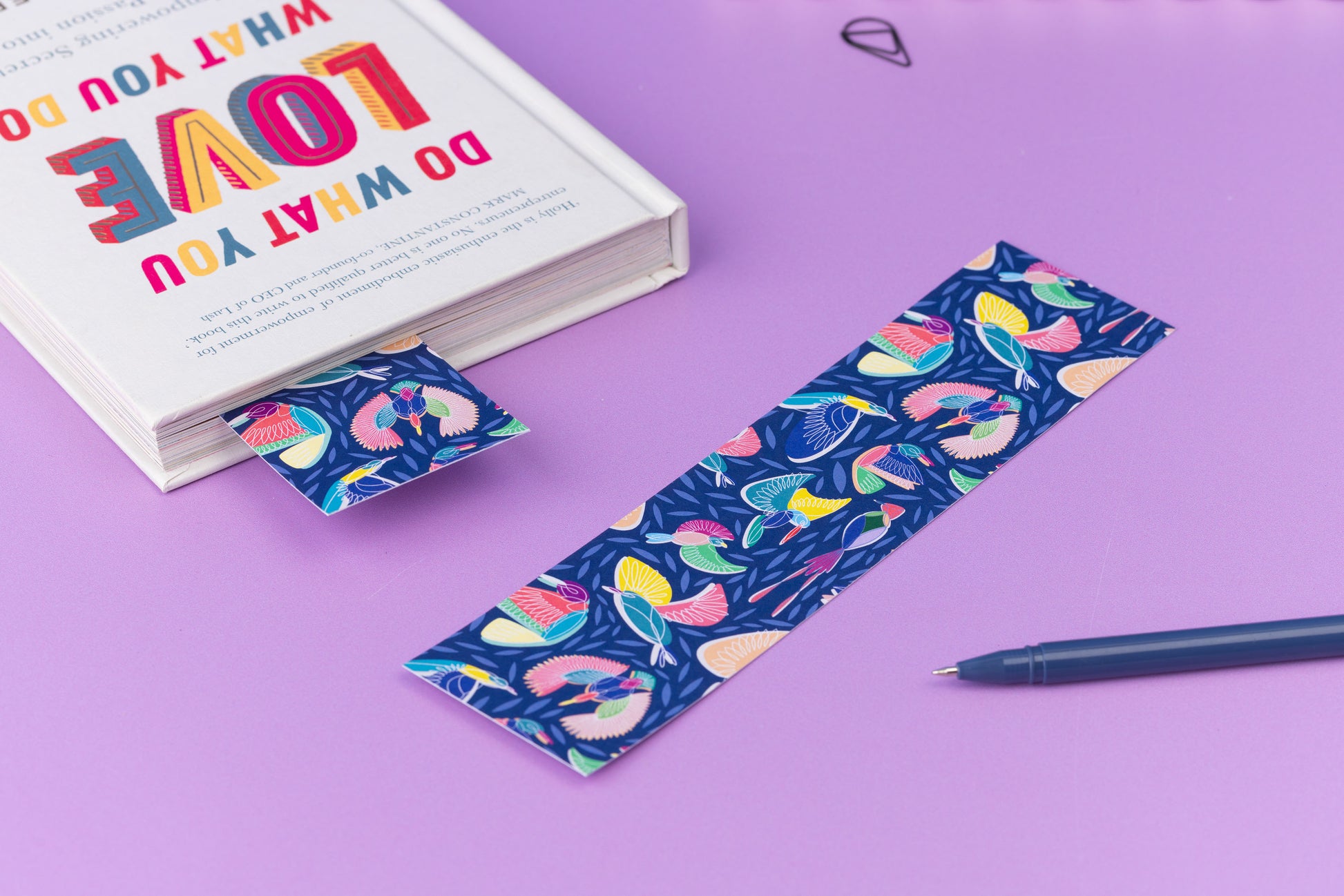 A Paradise double-sided bookmark is lying on a lilac desk with its full-colour side facing up.  To its left is a close book which has a Paradise bookmark sticking out of it, also full-colour side up.