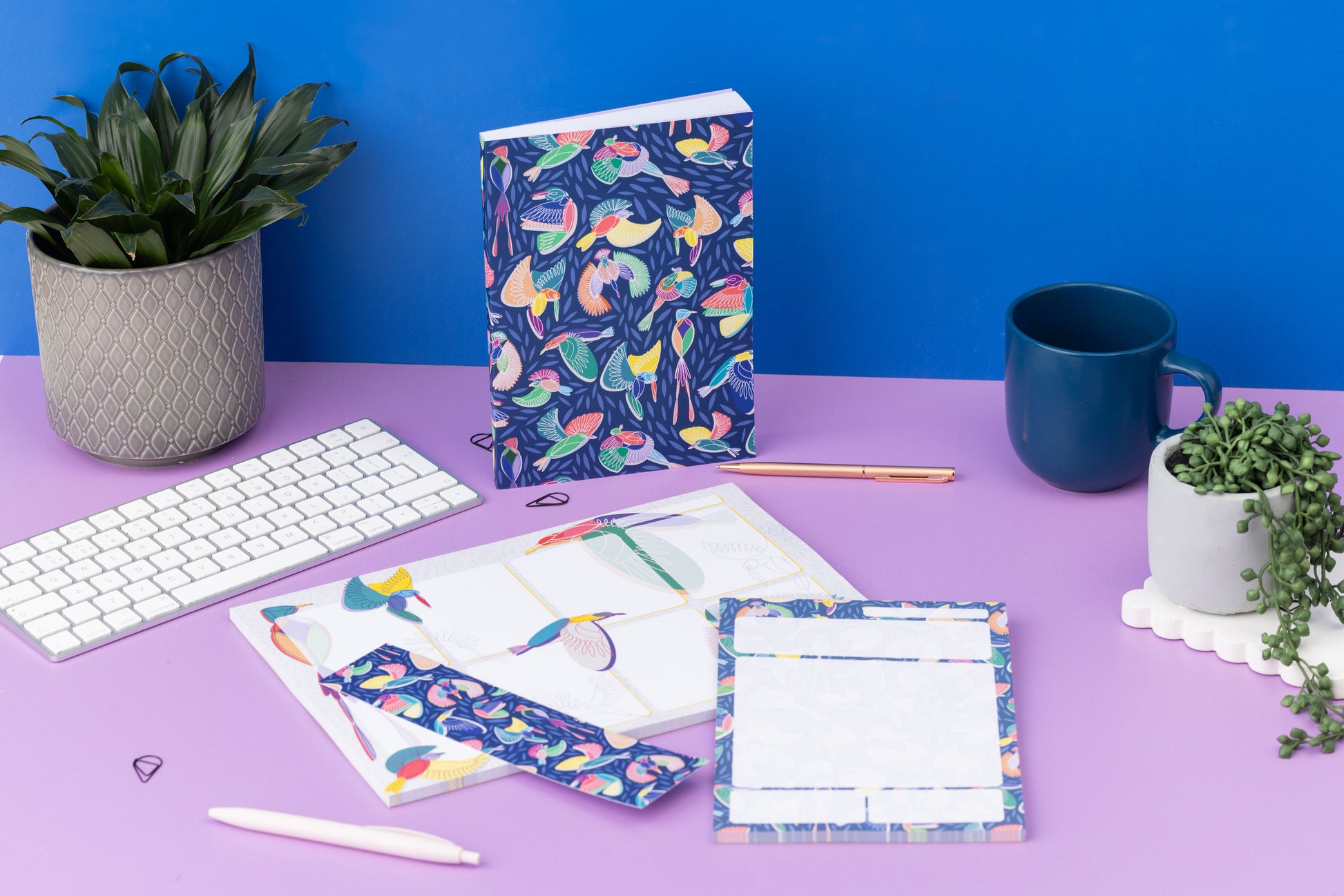Paradise Full collection - A4 Planner Pad, A5 Daily Planner Pad, Double-sided bookmark and A5 Dot Grid Notebook - are on a lilac desk with small plants, pens and a white keyboard around them.  The A5 Notebooks is upright.  There is a blue wall behind.
