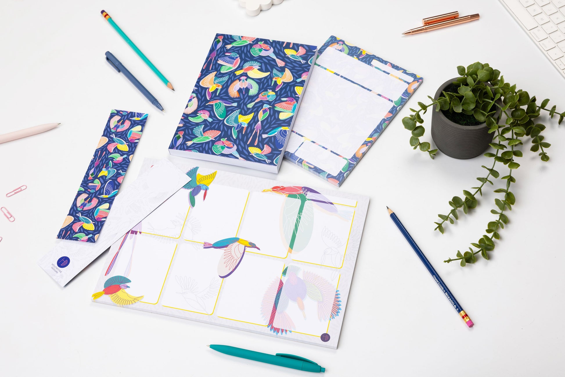 The Full Paradise collection - A4 Planner pad, A5 notebook, A5 Daily Planner Pad and 2 Double-side bookmarks (one side each) - are lying on a white desk surrounded by pens, paperclips and a small plant. 