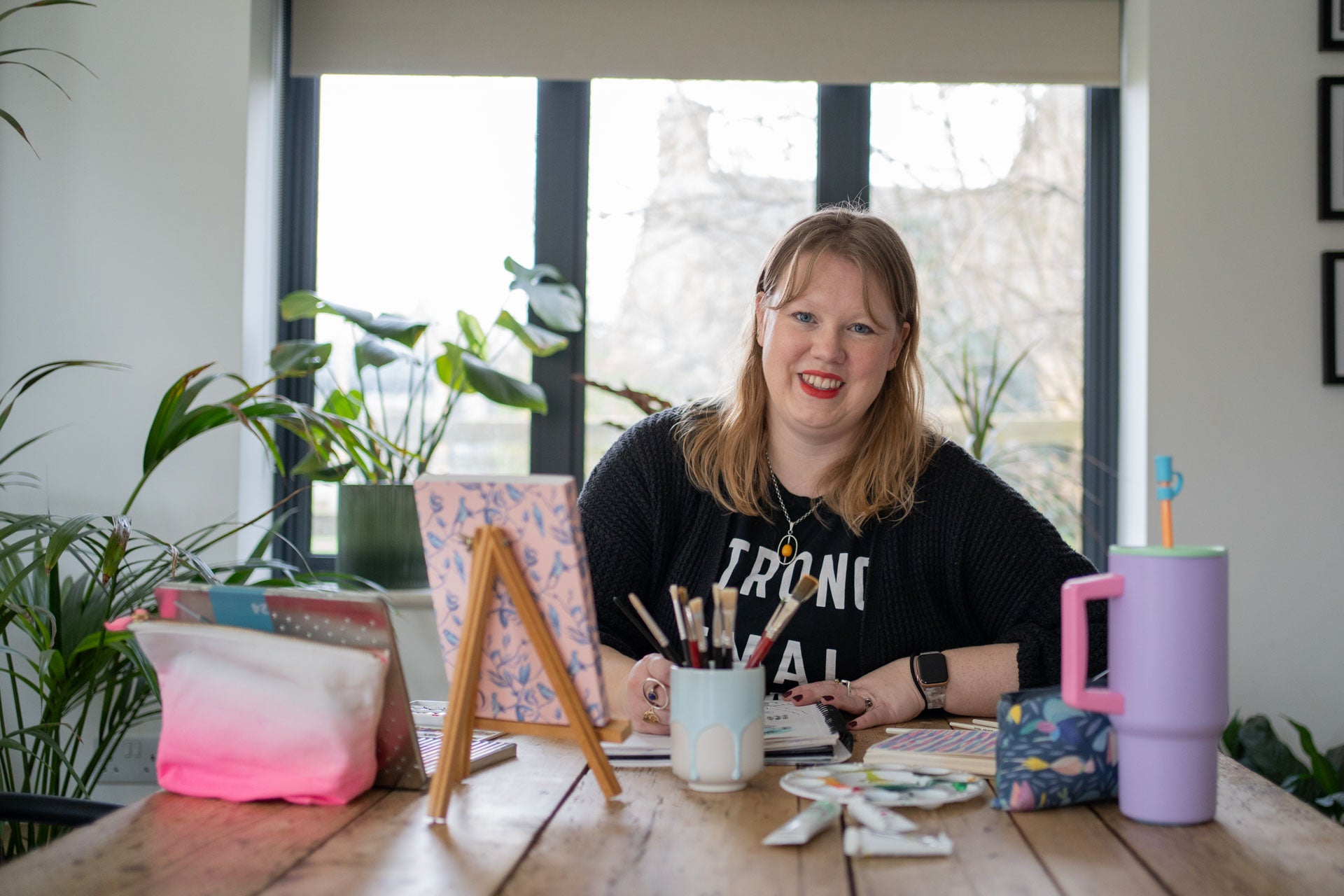 Rachel, a white woman in her 40s, is sitting at a table with art supplies and plants in a bright room.