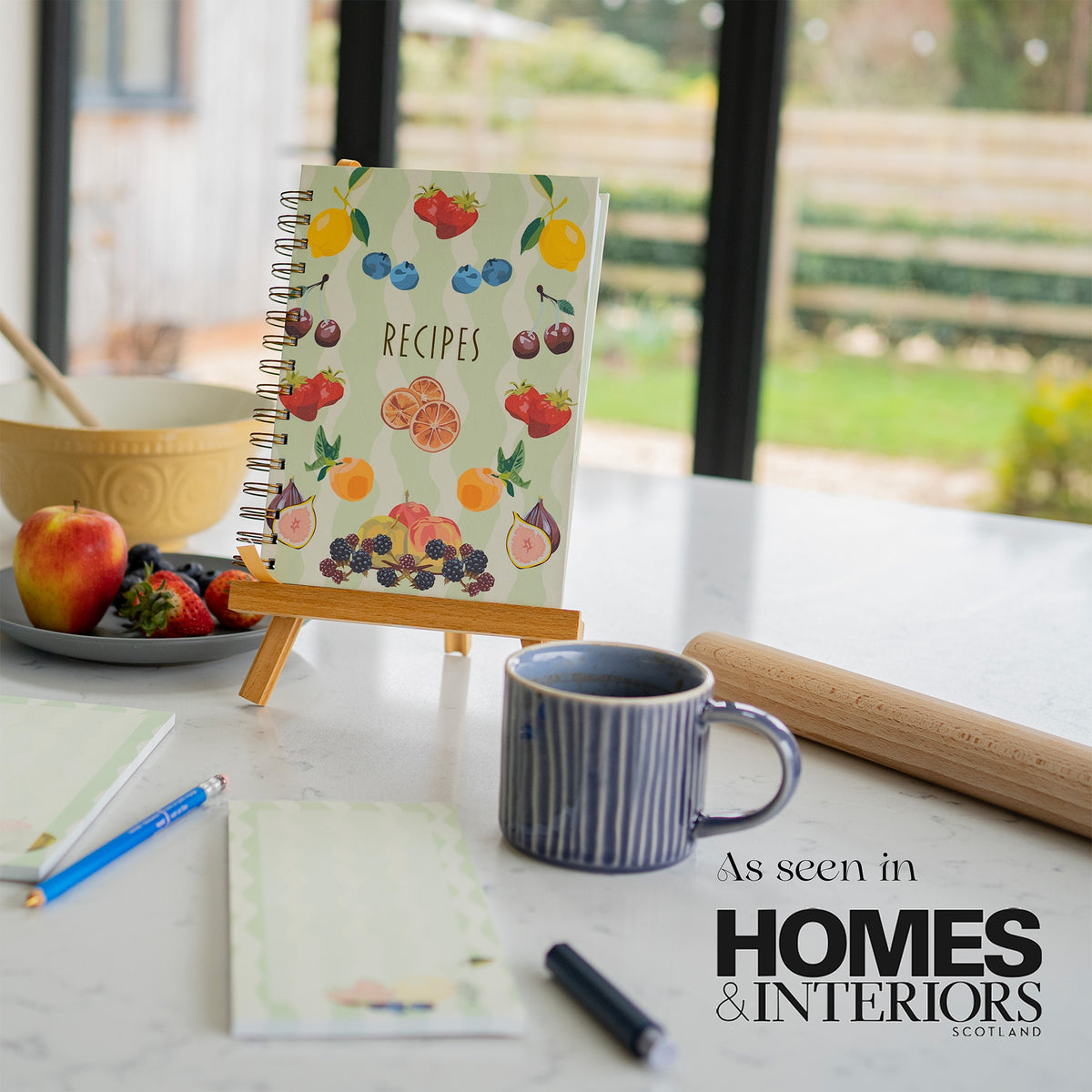 A spiral-bound recipe notebook with a colourful fruit illustration stands on a small wooden easel on a kitchen counter. The scene includes a blue striped mug, a rolling pin, fruit, matching notepads and pens, with a large window and outdoor view in the background. Overlayed text says as seen in Homes and Interiors Scotland. 